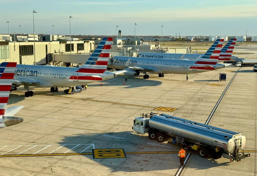 Several American Airlines jets parked at gates with jet bridges, ground crew and equipment on the tarmac.