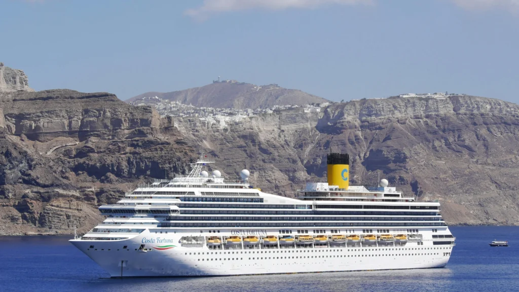 Costa Fortuna cruise ship with yellow funnel sits on calm blue water, rocky coastline and a hillside town in the distance.