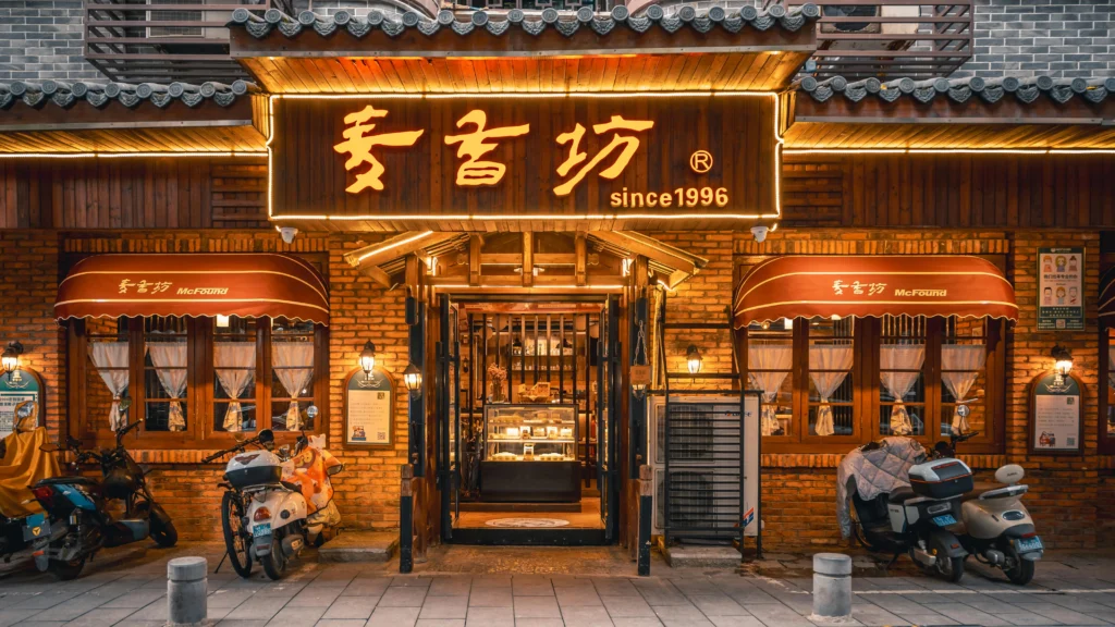 Brick storefront with warm wooden facade and illuminated Chinese sign reading 'since 1996'; two brown awnings, glass doors, and scooters parked at the entrance.