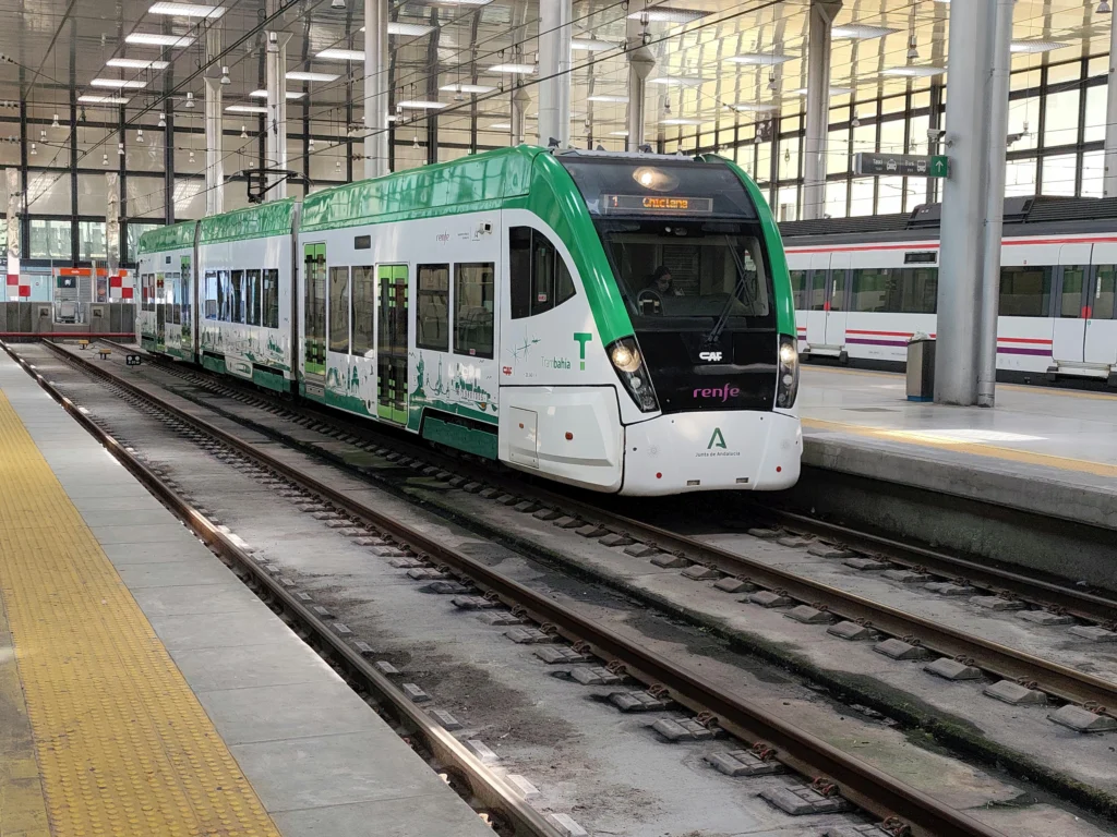 Green and white tram at a station platform with yellow tactile paving, and another train on the adjacent track visible in the background.