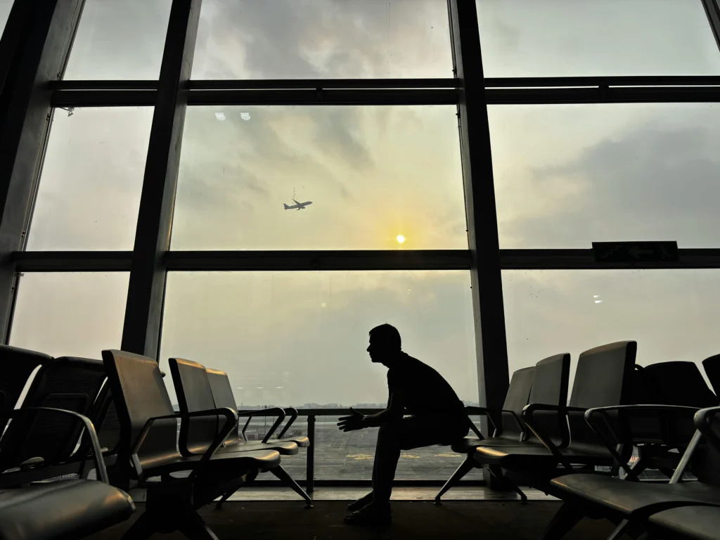 Silhouette of a person seated in an airport lounge, facing a large window as a plane takes off at sunset.