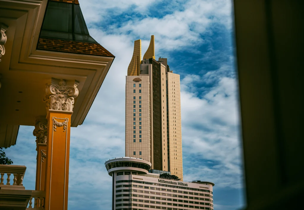 Cityscape featuring a modern Millennium Hilton hotel tower framed by a classical ornate balcony in the foreground against a blue sky.