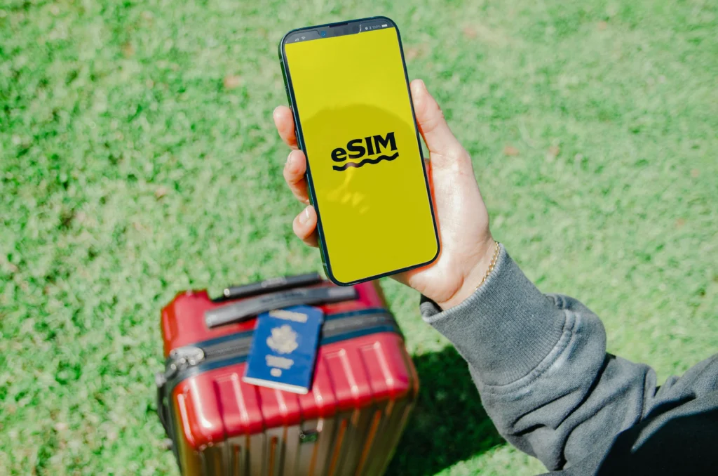 Smartphone displaying an eSIM screen while a traveler holds it near a red suitcase on green grass.