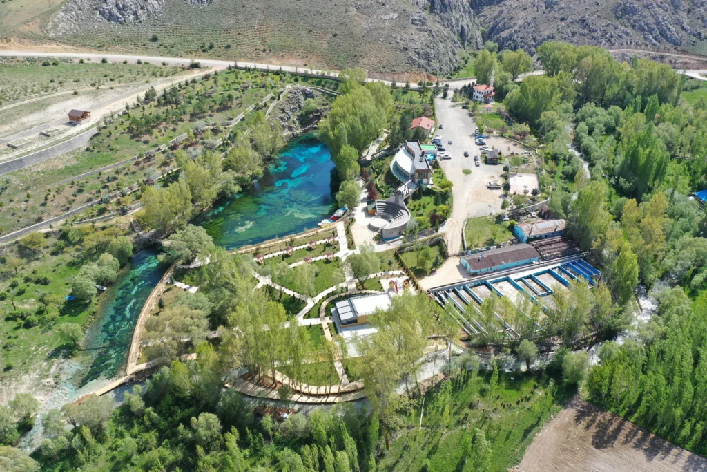 Aerial view of a park by a turquoise river, with green trees, paths, and buildings clustered around a central pool area near a hillside.