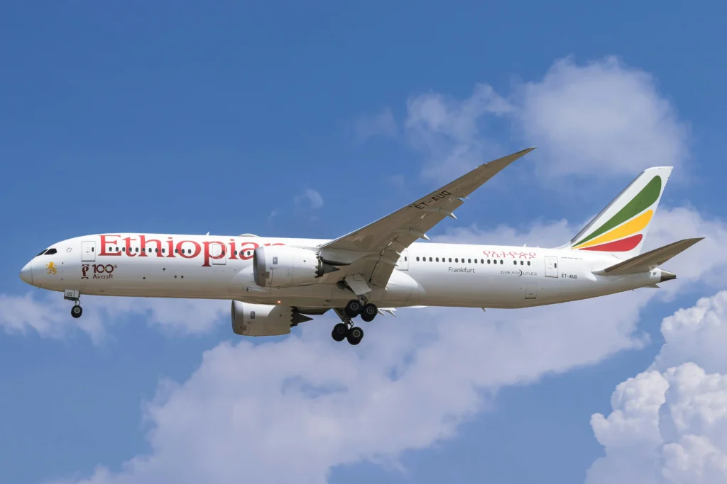 Ethiopian Airlines aircraft in flight with landing gear down, white fuselage and colorful tail against a blue sky with clouds.