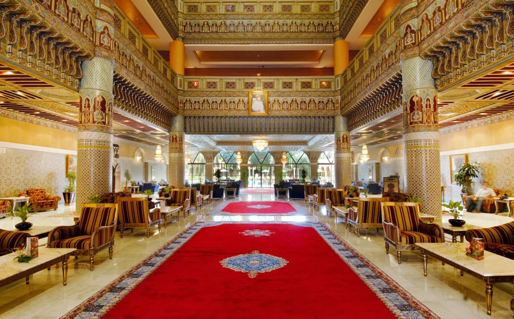 Opulent hotel lobby with ornate gold-and-ceramic columns, arches, and a long red carpet running through the center.