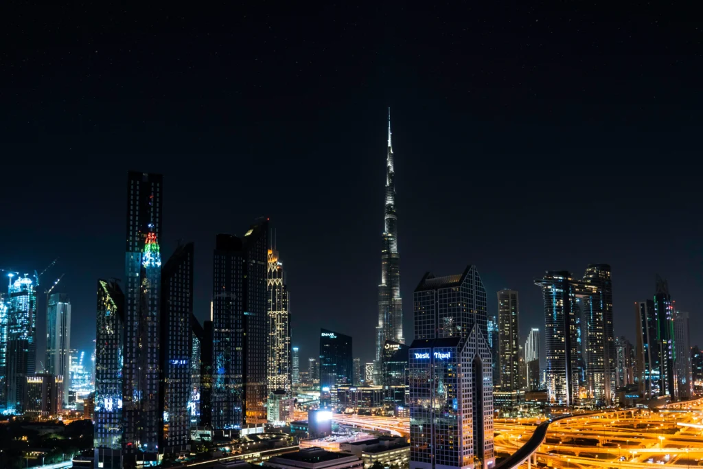 Nighttime city skyline with the tall illuminated Burj Khalifa rising above glass skyscrapers in Dubai.