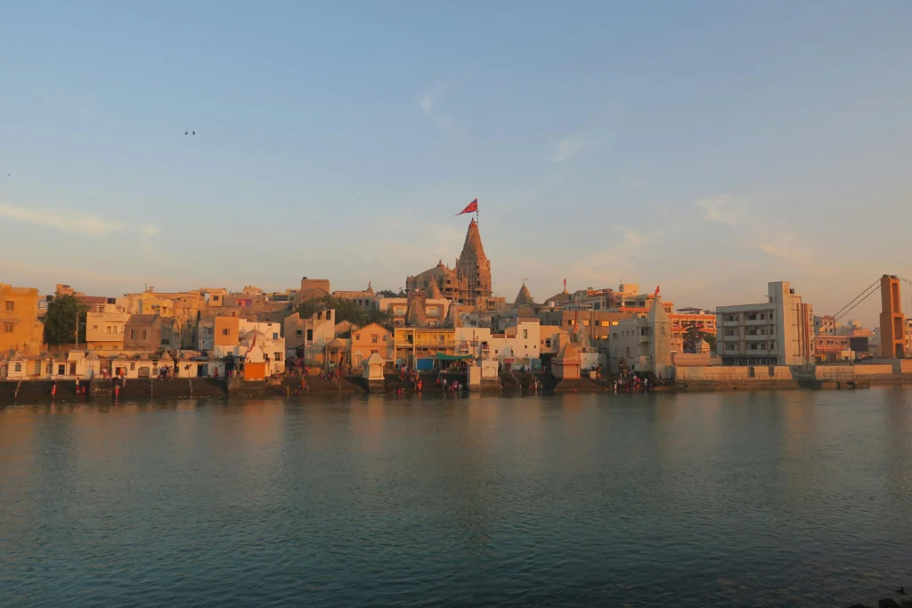Riverside town at sunset with a hilltop temple featuring a red flag overlooking colorful buildings along the water.