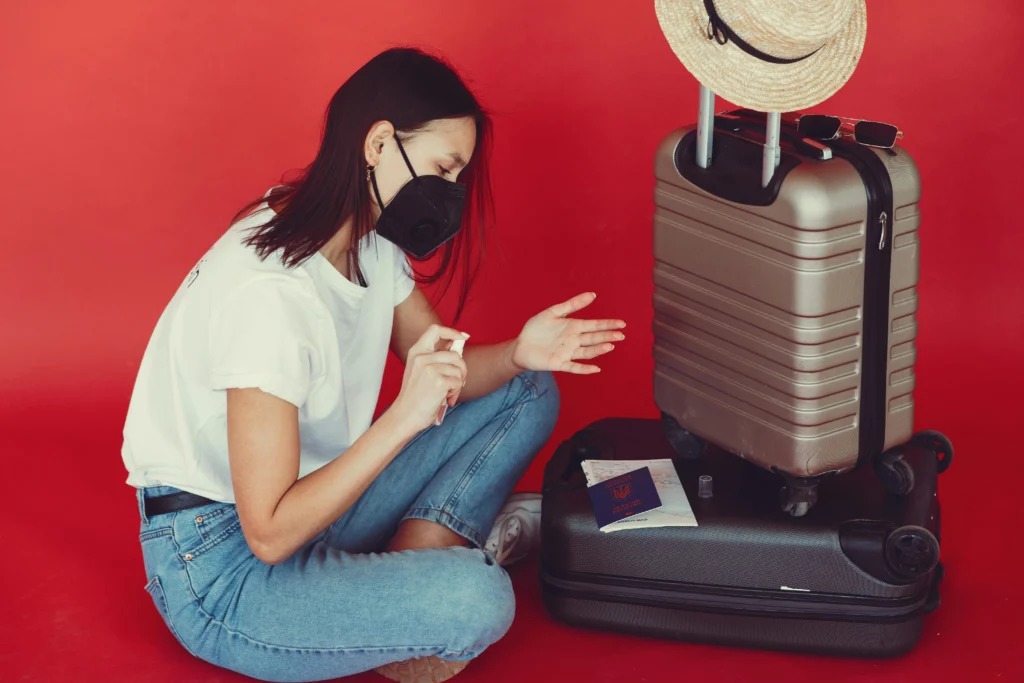 Woman in a black face mask sits next to a silver suitcase on a red background, with a straw hat and sunglasses resting on top.