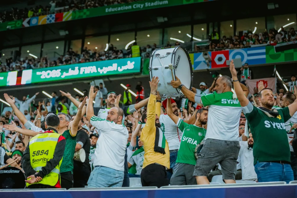 Fans in green and white jerseys celebrate in a stadium, a large drum raised above the crowd amid cheering spectators.