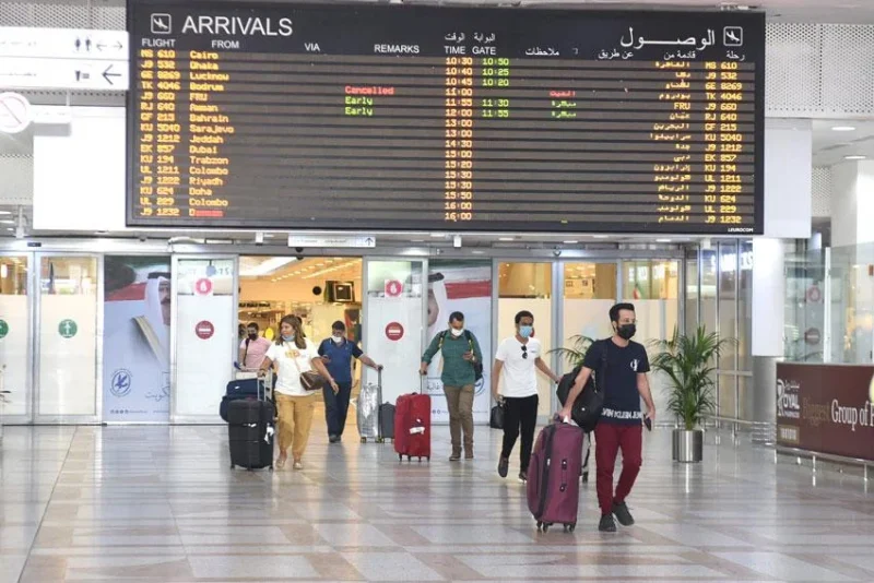 Airport arrivals hall with a large flight information board overhead; travelers with rolling luggage walking through.
