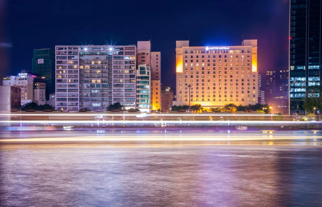 Night city skyline with lit high-rise buildings and light trails across a river in the foreground.