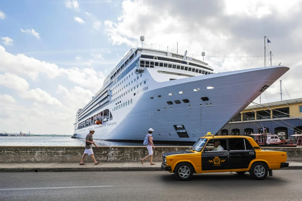 Large white cruise ship docked at a harbor, with a yellow taxi and two pedestrians walking along the quay on a sunny day.