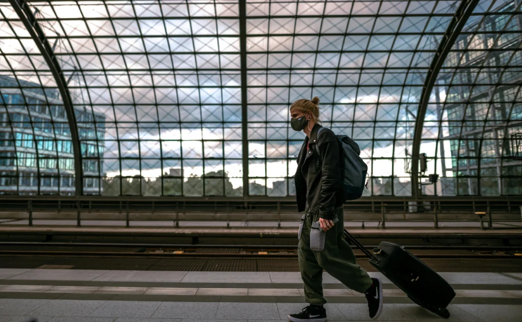 Traveler wearing a face mask walks with a rolling suitcase along a modern glass-covered train station platform.