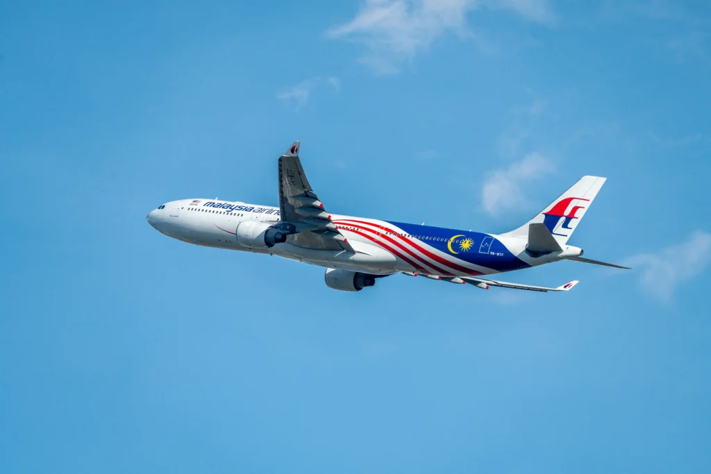 Malaysia Airlines jet in flight across a clear blue sky, tail logo visible on the right.