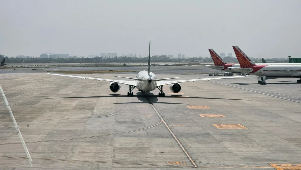 Front view of a commercial airliner taxiing on the airport tarmac, with two red-tailed planes parked in the background on the right.