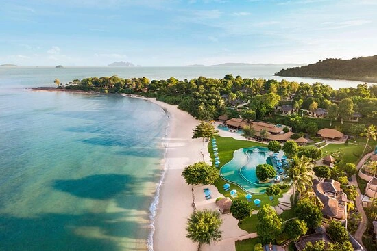 Aerial view of a tropical resort with a curved pool, white sand beach, and palm trees along a turquoise sea.