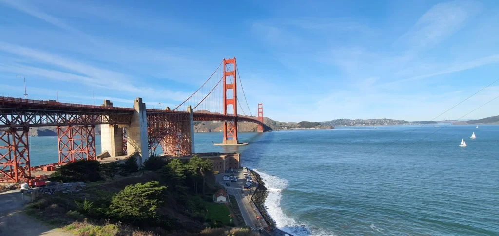 Red Golden Gate Bridge spanning a blue bay on a sunny day, with sailboats and hills in the distance.