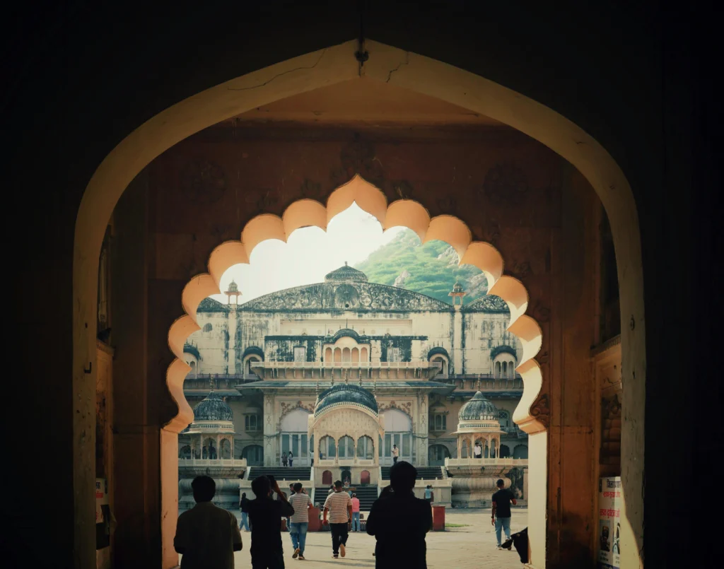 Visitors walk through a dark arch into a sunlit Indian palace courtyard with scalloped arches and domed pavilions in the background.