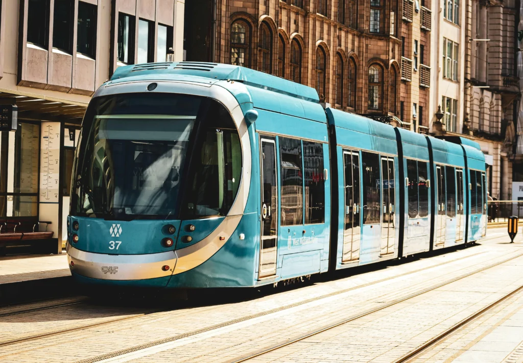 Front and side view of a teal-blue tram stopped at a city platform with brick buildings in the background.