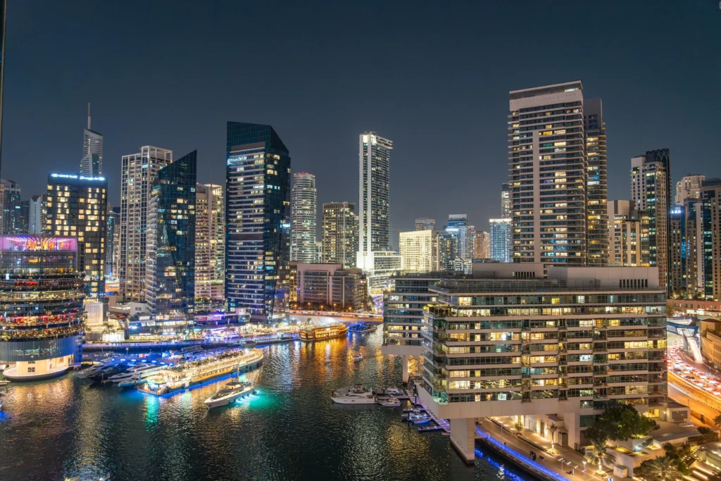 Nighttime city skyline with illuminated skyscrapers along a busy marina and calm water.