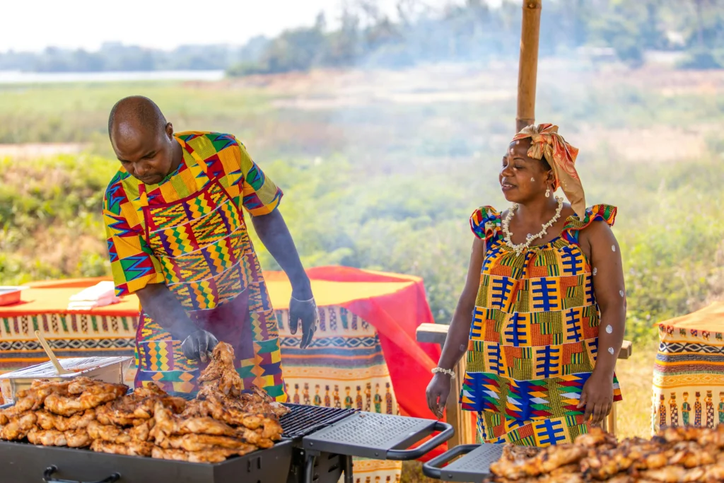 Man and woman in vibrant, patterned outfits grilling skewered meat outdoors at an event.