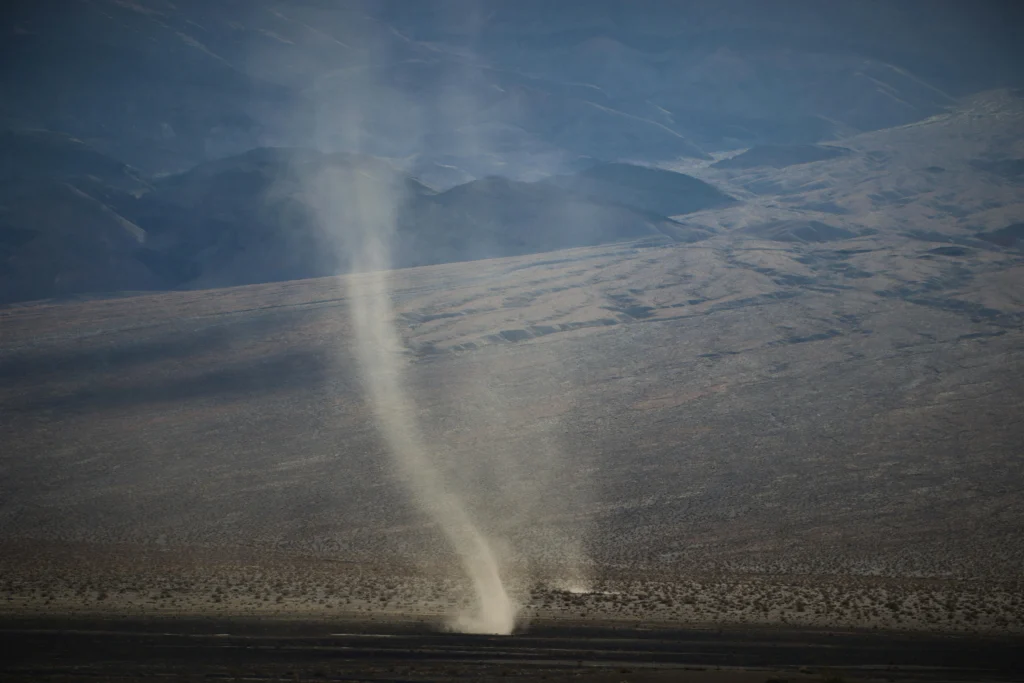 Desert landscape with a pale vertical sunbeam or dust column rising from the ground into a hazy blue sky, mountains in the distance.