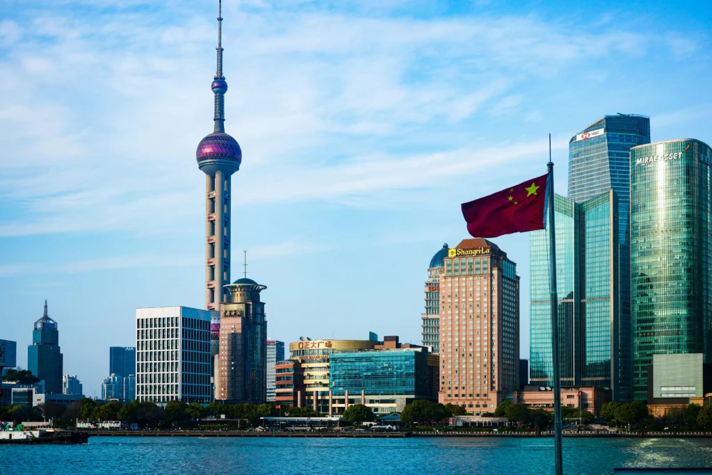 Shanghai skyline with the Oriental Pearl Tower beside glass skyscrapers and a waving Chinese flag by the river foreground.