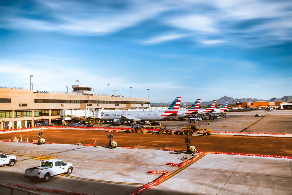 Airfield at a sunny airport with several American Airlines planes at gates and ground support vehicles nearby.