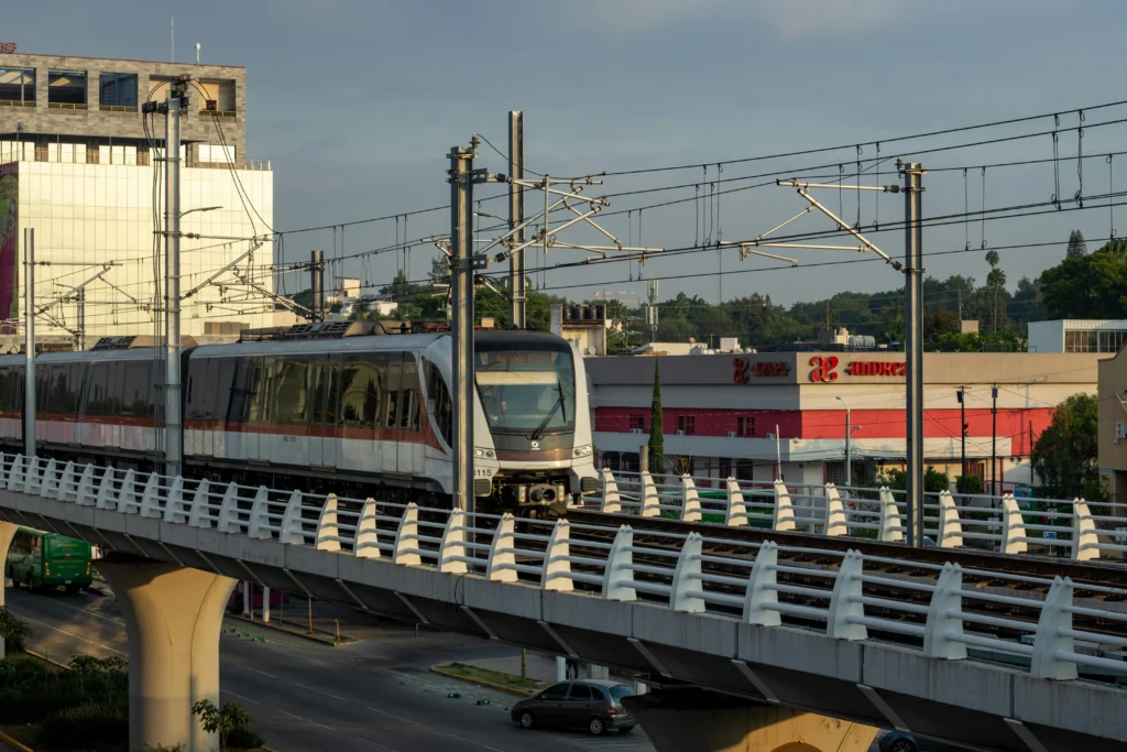 Passenger train on an elevated track with overhead power lines against a city background.