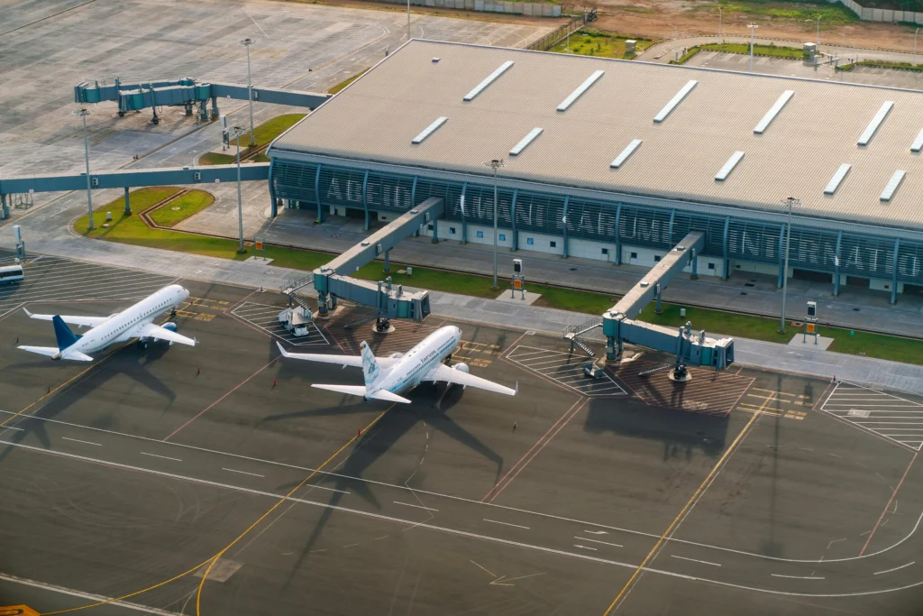 Aerial view of a modern airport terminal with jet bridges and parked airplanes at gates on the tarmac in front of the building, with taxiways nearby.