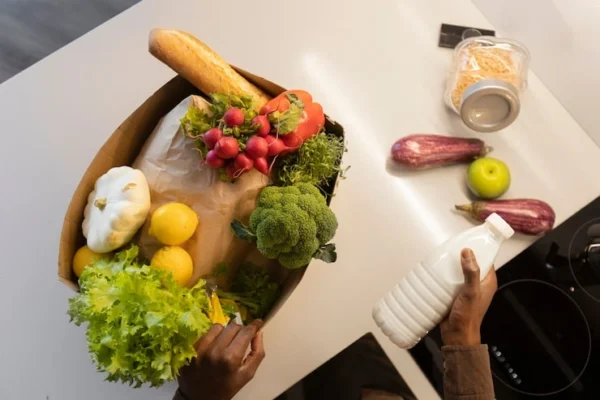 Person holds a basket of fresh produce (lettuce, broccoli, squash, radishes, lemons, a baguette) on a kitchen counter with other items nearby (milk bottle, apples, eggplants).