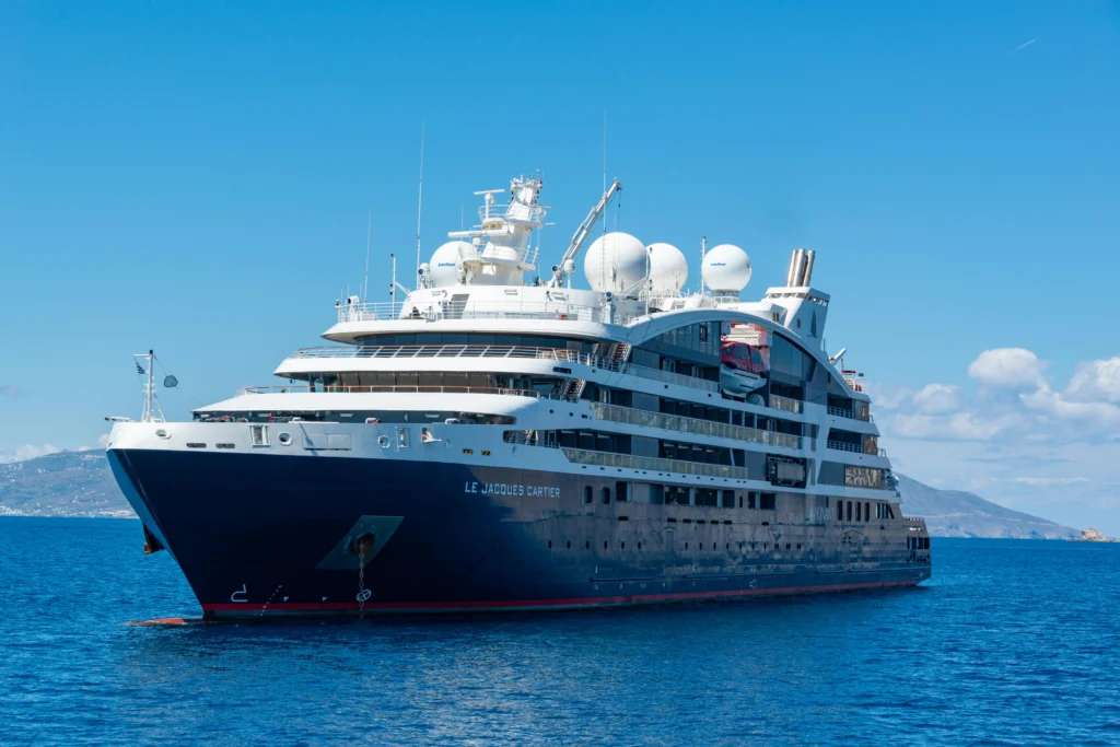 Luxury cruise ship anchored on calm blue sea under a clear sky, with multiple decks and satellite domes visible on top.