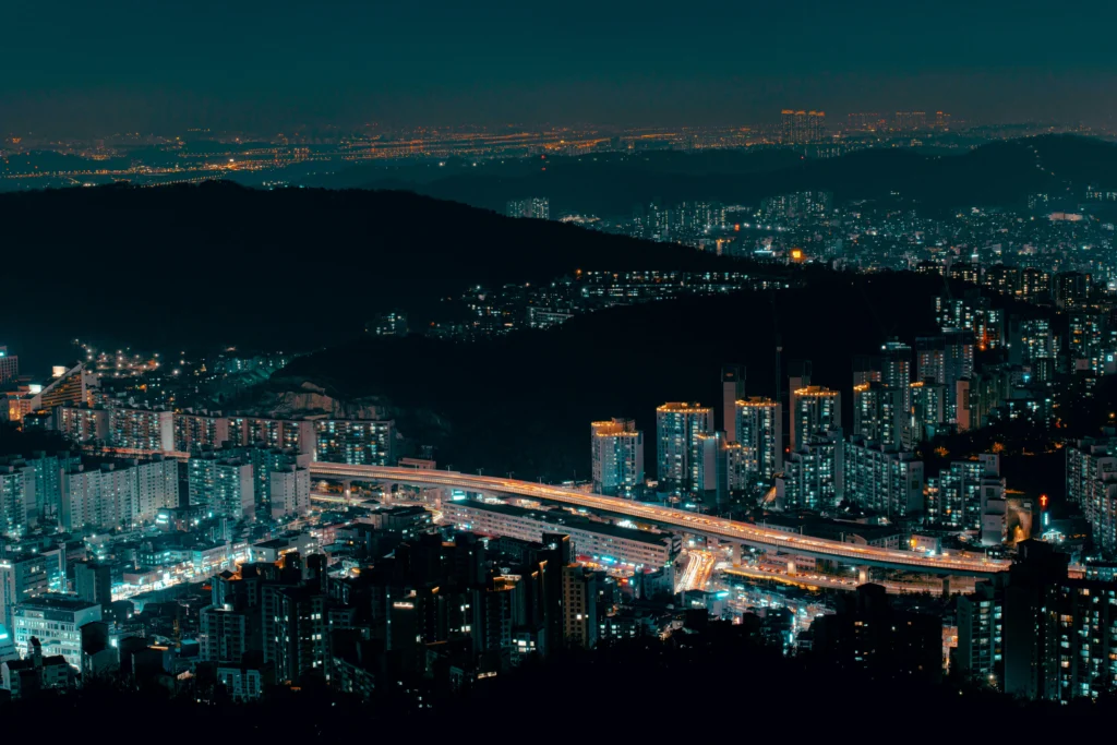 Nighttime cityscape: densely packed buildings glowing with street and window lights, hills in the foreground and a lit highway below.