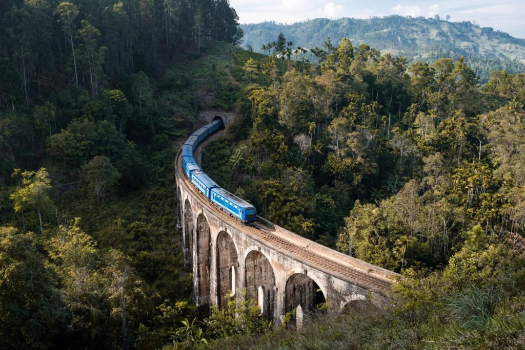 Blue passenger train winding along a stone viaduct through dense tropical forest. An arched bridge crosses a deep valley surrounded by trees.