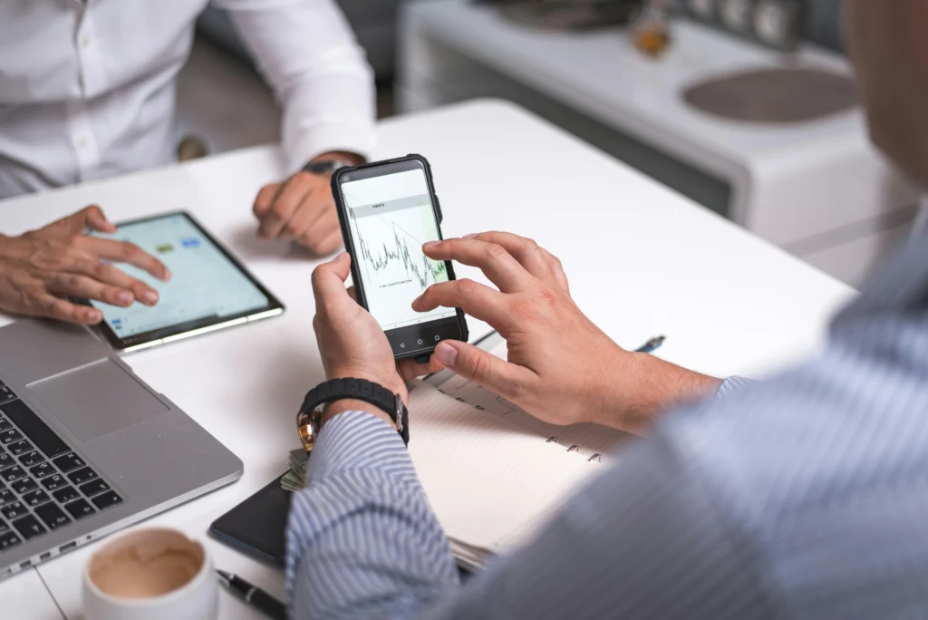 Hands holding a smartphone showing a financial chart while a tablet and laptop sit on a white desk in an office setting.
