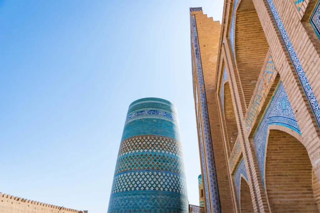 Tall turquoise tiled minaret leaning beside a brick arcade with blue tile mosaics under a clear blue sky