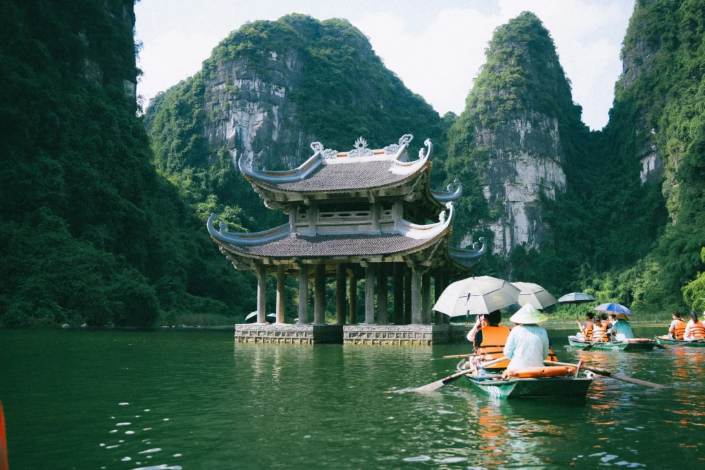 Traditional Chinese pavilion on stone pillars in a calm river, with lush limestone karst cliffs rising behind it and boats nearby with umbrellas.