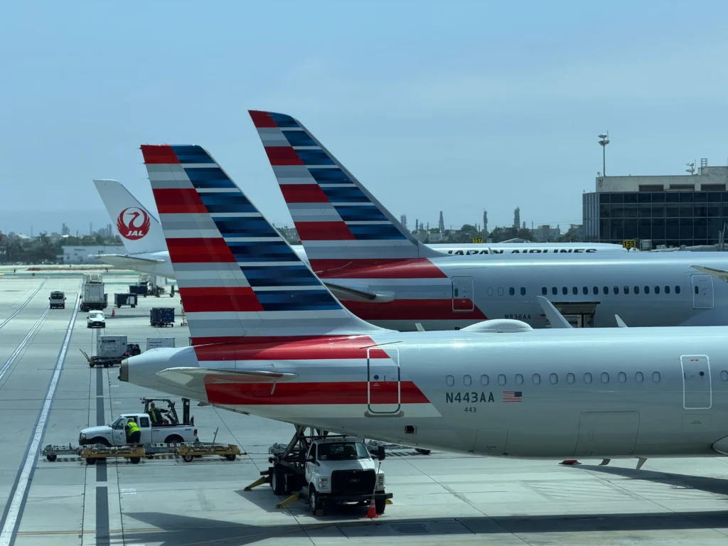 Rear view of two American Airlines aircraft tails with red, white, and blue stripes at the gate, with ground crew and tarmac equipment visible below.