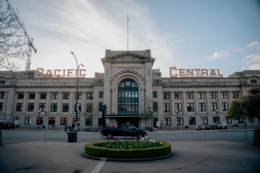 Pacific Central Station facade with orange sign, central arch, flagpole, and a circular flower bed in front.