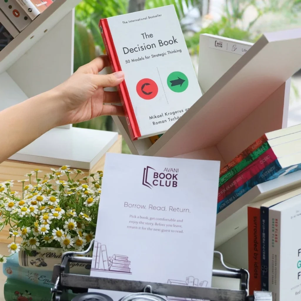 Hand holding 'The Decision Book' with red spine, about to place it in a book club display shelf with daisies nearby.