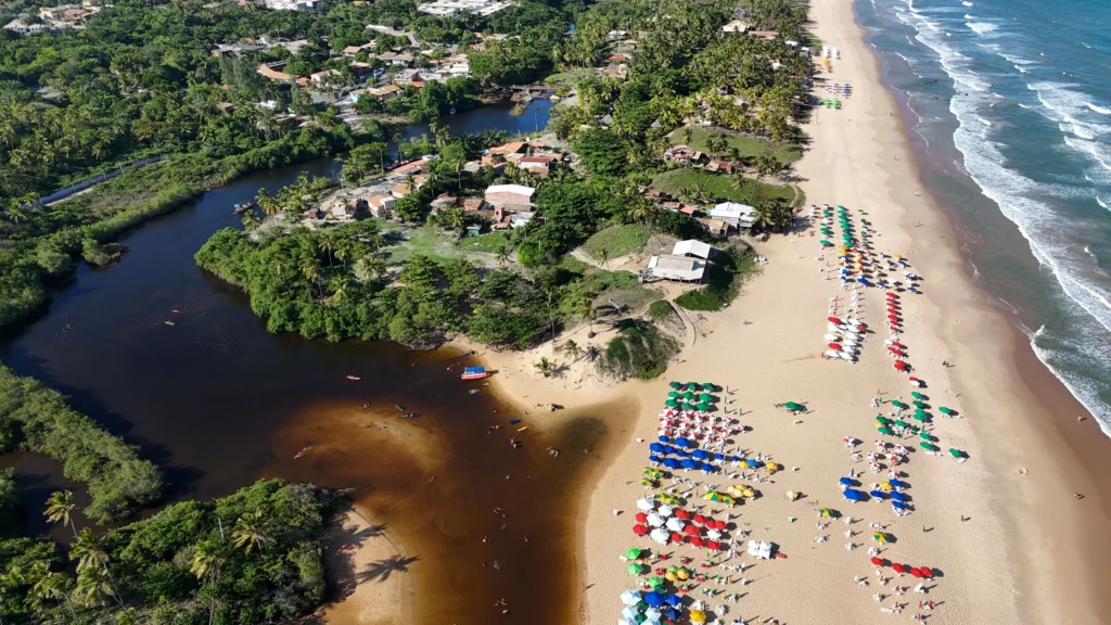 Aerial view of a tropical beach with rows of colorful umbrellas and sunbathers along the shore, the blue ocean to the right and a river on the left.