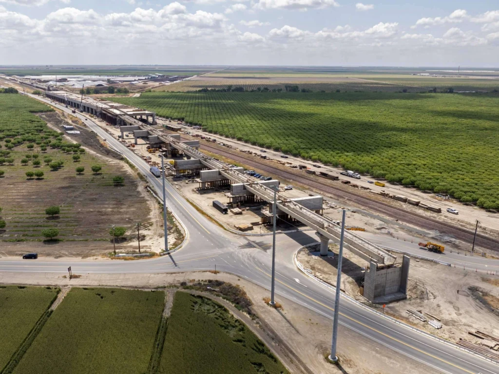 Aerial view of an industrial construction site with an elevated conveyor system beside railway tracks and surrounding fields.