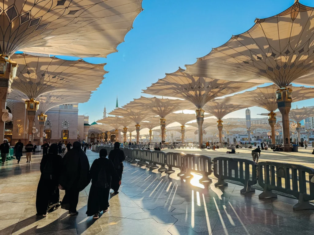 Wide courtyard under large white fabric canopies with geometric patterns; people in black abayas walking and shadows on the polished floor mix with golden lanterns nearby.