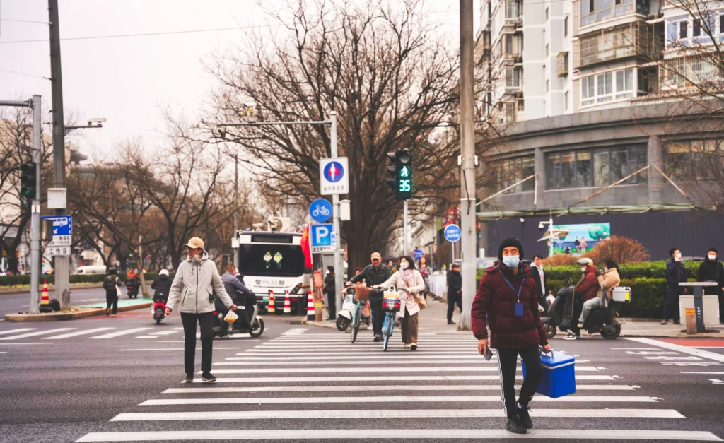 Pedestrians and cyclists cross a busy city crosswalk, many wearing masks, with buildings and traffic signals in the background.