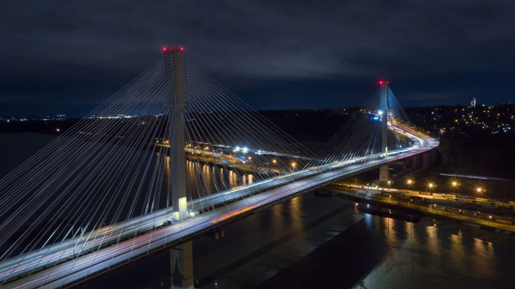 Nighttime aerial view of a cable-stayed bridge with tall pylons and many white suspenders, illuminated over a dark river and city lights in the distance.