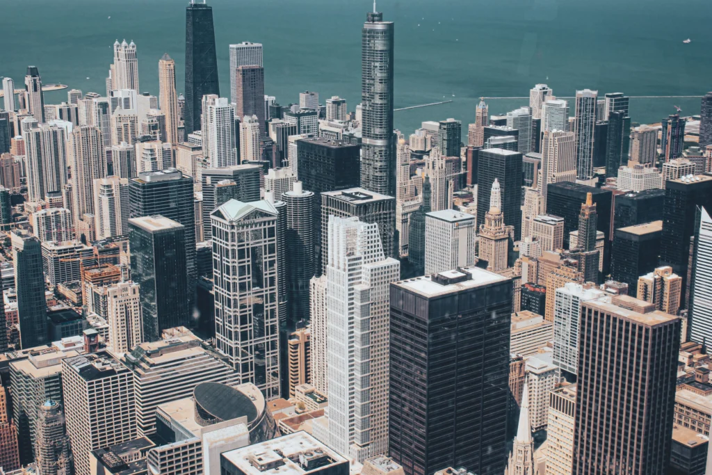 Aerial view of a dense downtown skyline with numerous skyscrapers along a blue-green waterfront pond of water behind them.