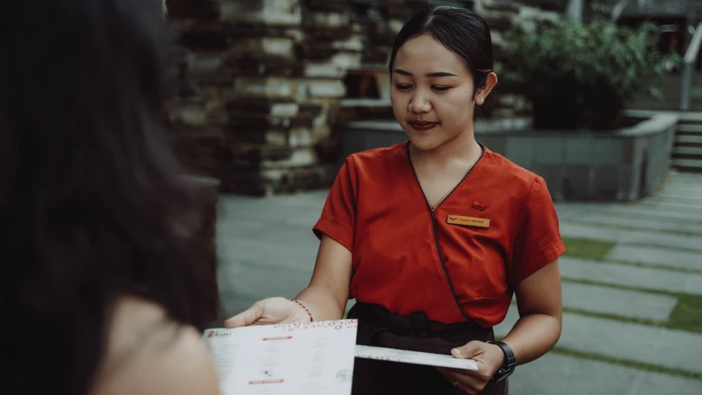 Server in an orange uniform with a name badge 'YULITA-TRAINEE' hands a paper menu to a customer at an outdoor table.