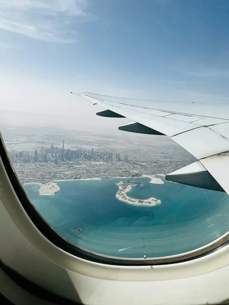 View from a passenger window showing an airplane wing over a coastal city with a dense skyline and blue water below.