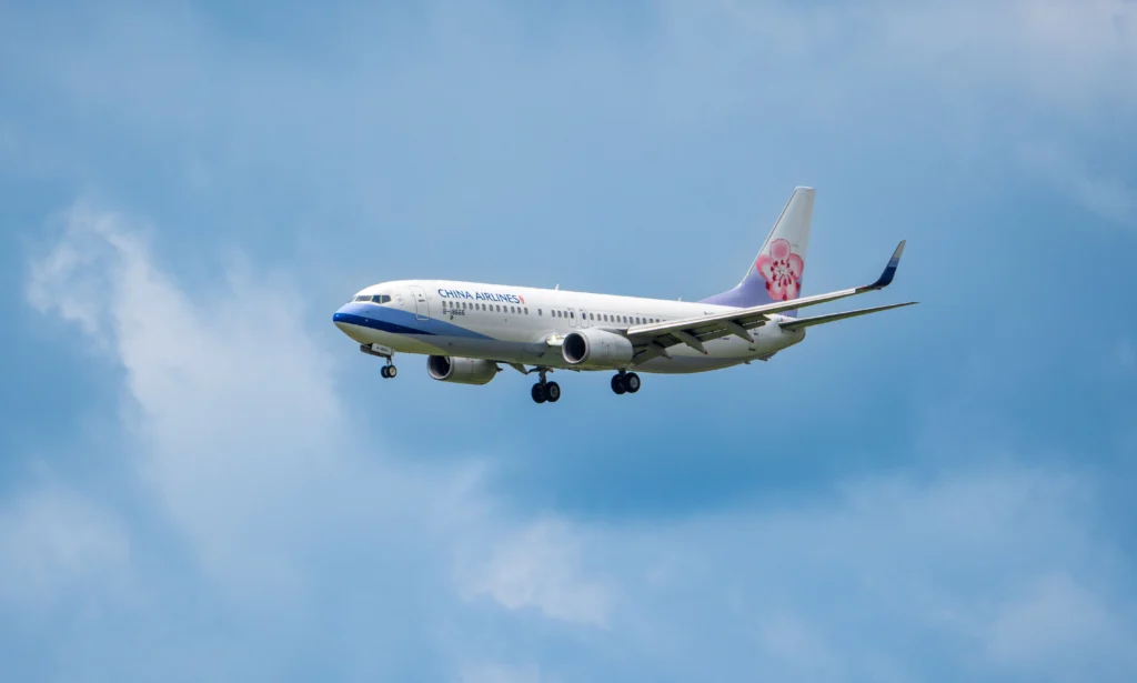 Commercial airliner in flight against a blue sky, logoed with a pink flower on the tail (China Airlines).